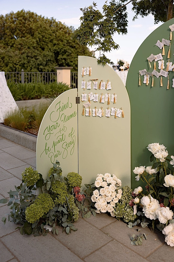 Wedding seating chart with arched seating chart board and hanging tassel escort cards, framed by white roses, peonies, and hydrangeas on a garden patio
