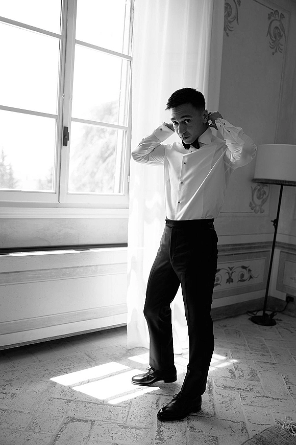 Groom portrait in a white dress shirt and black bow tie, standing by a large window with natural light and patterned wall behind