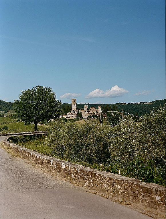 Wedding venue landscape with a stone wall leading to rustic stone buildings and a bell tower, set against rolling hills under blue sky clouds