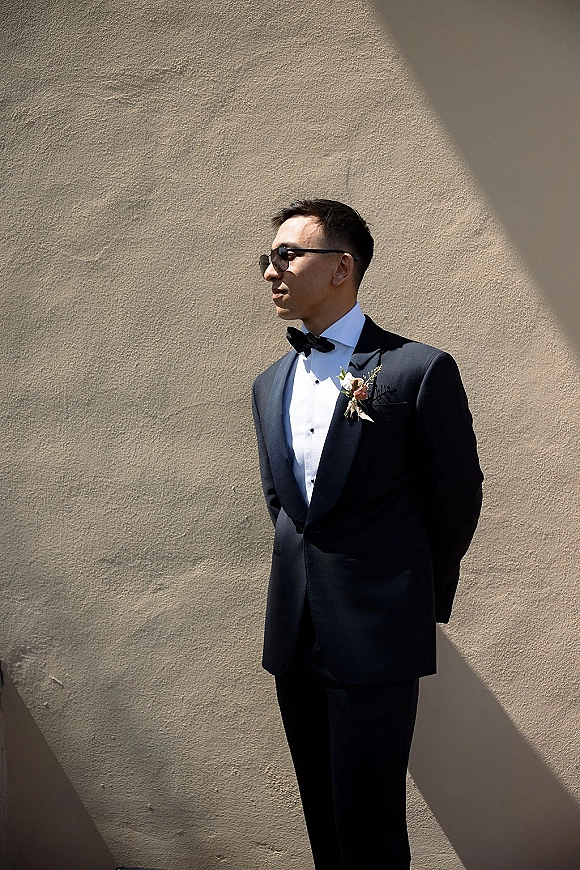 Groom portrait in a black tuxedo with bow tie and boutonniere, wearing sunglasses by a stucco wall with sharp sunlight shadows