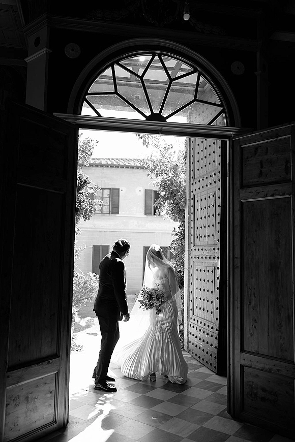 Wedding couple portrait in black and white, bride in veil holding bouquet beside groom in suit at arched wooden doorway with shadows