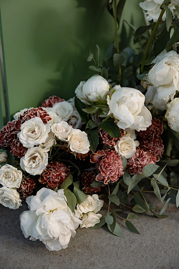 Wedding florals featuring white peonies and roses with rust carnations and eucalyptus greenery, arranged on stone before a green wall