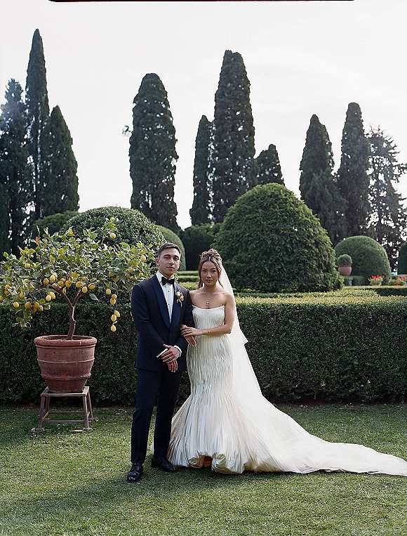 Couple portrait of bride in strapless gown and veil holding groom’s arm, tuxedo with boutonniere, in a formal garden with cypress trees