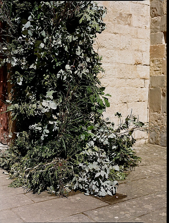 Ceremony floral installation with greenery ceremony backdrop of eucalyptus and branches against a stone wall in an outdoor courtyard