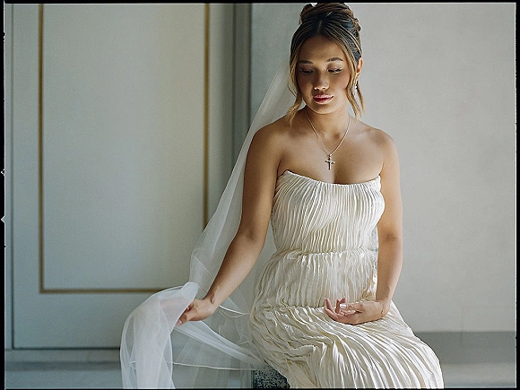 Bridal portrait of a bride sitting portrait in a strapless pleated gown, holding her veil and looking down by a paneled wall in soft window light