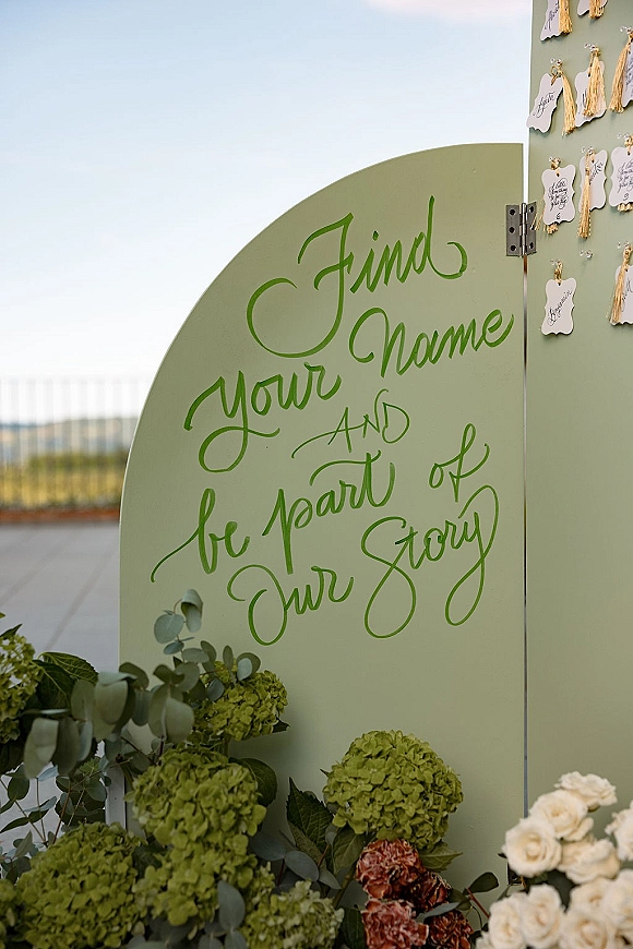 Wedding escort display and wedding seating chart on a mint green calligraphy board with hanging tassel cards and hydrangeas on a rooftop terrace