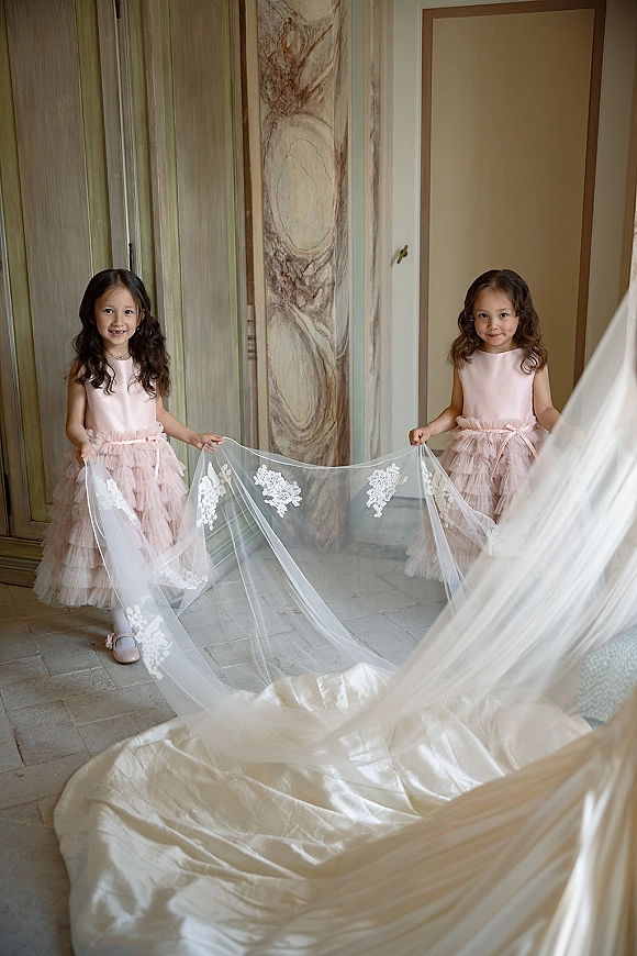 Flower girls in blush pink tulle dresses hold the bride’s lace veil and train in an indoor hallway with marble floors and paneled doors