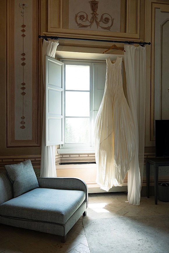 Wedding dress hanging by window in natural light on a hanger from a curtain rod, with curtains and a chaise lounge behind
