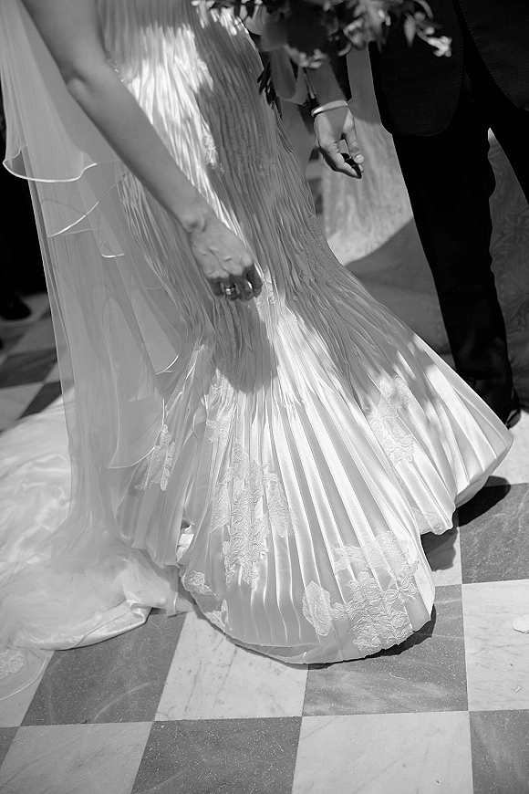 Wedding dress detail with pleated wedding dress skirt, lace appliqué hem and veil train flowing over a checkered marble floor indoors