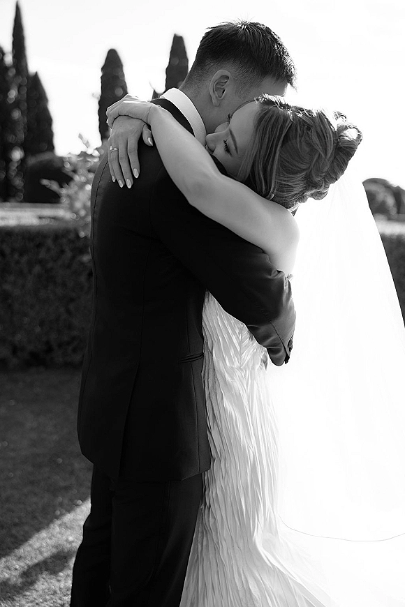 Wedding kiss portrait of bride and groom kiss in a garden, her long veil flowing behind a strapless dress as he holds her in a tuxedo