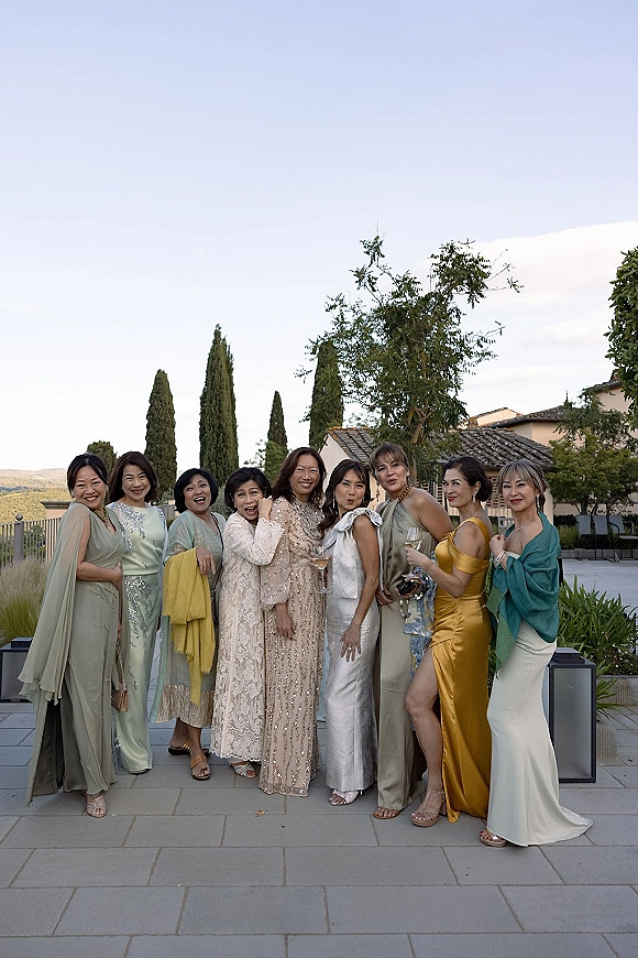 Bridesmaids group photo with a champagne toast, women in neutral dresses and shawls holding flutes on a stone patio with greenery