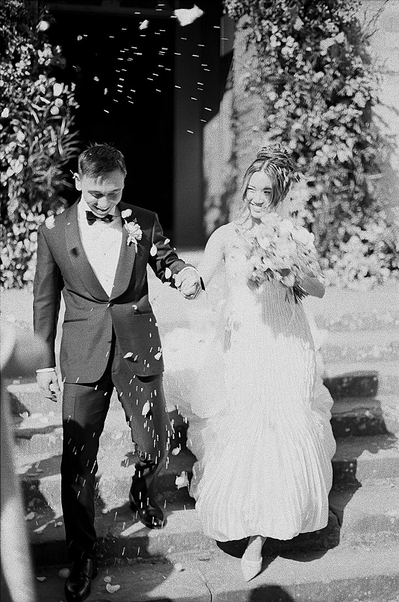 Wedding recessional as newlyweds walk out hand in hand, bride holding bouquet, petals in air on stone steps by a floral wall entrance