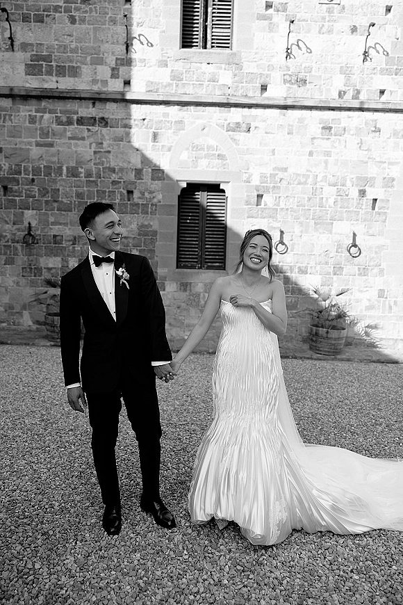 Couple portrait of bride and groom holding hands in a gravel courtyard, her strapless mermaid gown with long train beside a stone facade