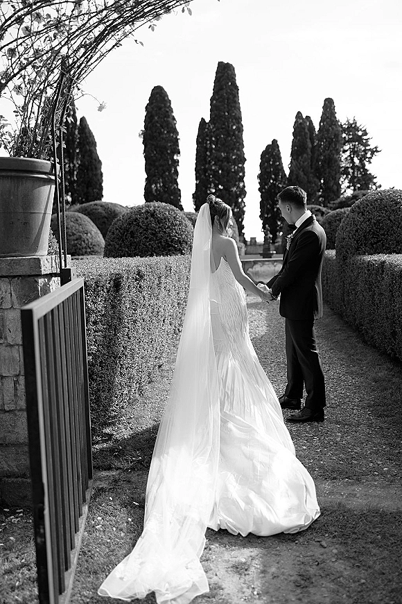 Couple portrait in black and white of bride and groom holding hands from behind, veil and train flowing along a tree-lined garden path