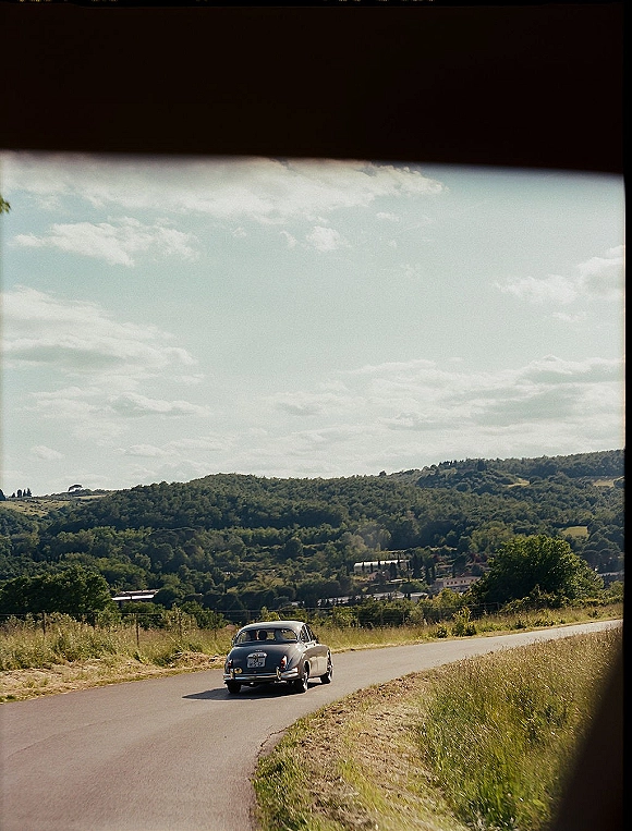 Wedding getaway car, a vintage car driving down a country road past grassy fields and rolling hills under a cloudy sky