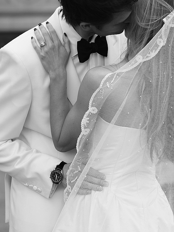 Wedding kiss close up of bride and groom kiss, lace-trim veil framing her ringed hand on his shoulder, blurred steps behind