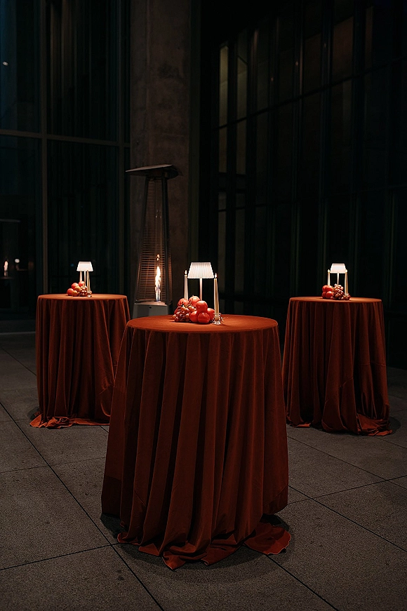 Cocktail hour decor with wedding cocktail tables draped in rust linens, topped with table lamps and a fruit centerpiece beside candles by a glass wall
