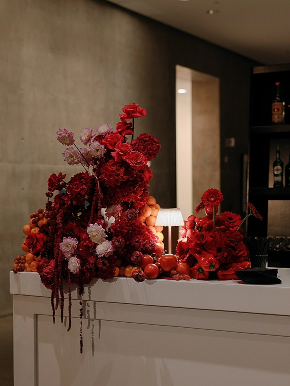 Reception bar decor with wedding bar styling, featuring cascading red roses, dahlias, amaranthus, and fruit on a white counter by liquor shelves