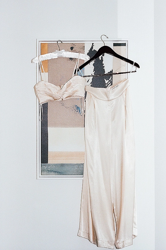 Wedding dress in satin slip style hanging on dress hangers against a white wall, with framed artwork nearby for minimalist detail