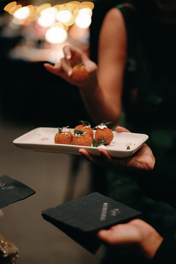 Wedding cocktail hour passed appetizers on a rectangular tray with herb garnish and cocktail napkins, set against warm string lights bokeh