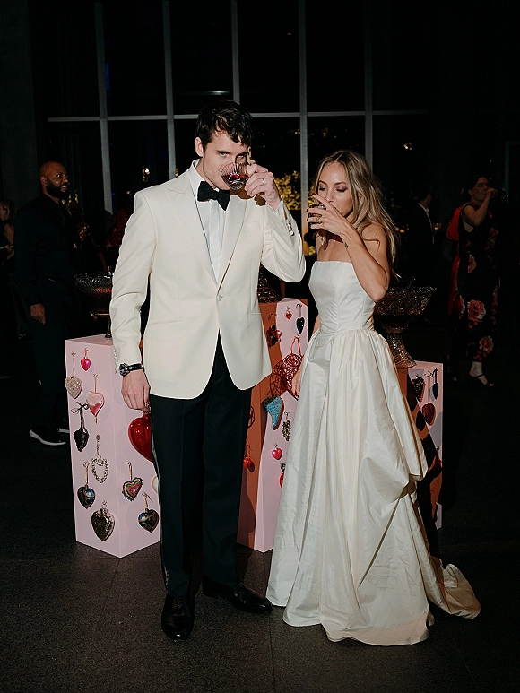 Reception toast moment as the bride and groom drinking cocktails, her strapless gown and his white jacket, by large windows at night