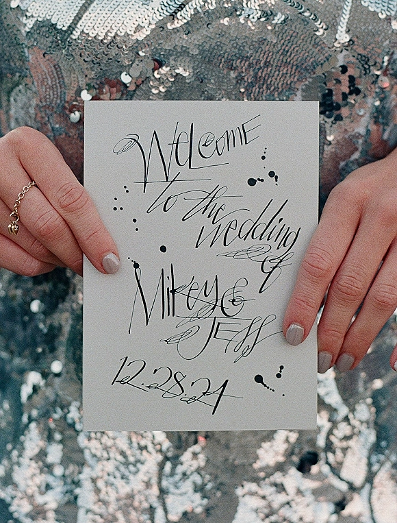 Wedding welcome sign with calligraphy wedding welcome sign lettering on white cardstock, held by manicured hand over silver sequin fabric background