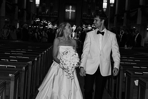 Wedding recessional as bride and groom walk the church aisle holding hands, bride with cascading bouquet and strapless dress, guests in pews behind