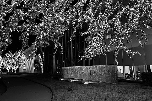 Museum wedding venue exterior at night with string light canopy over illuminated trees, stone sign by sidewalk, and glowing windows