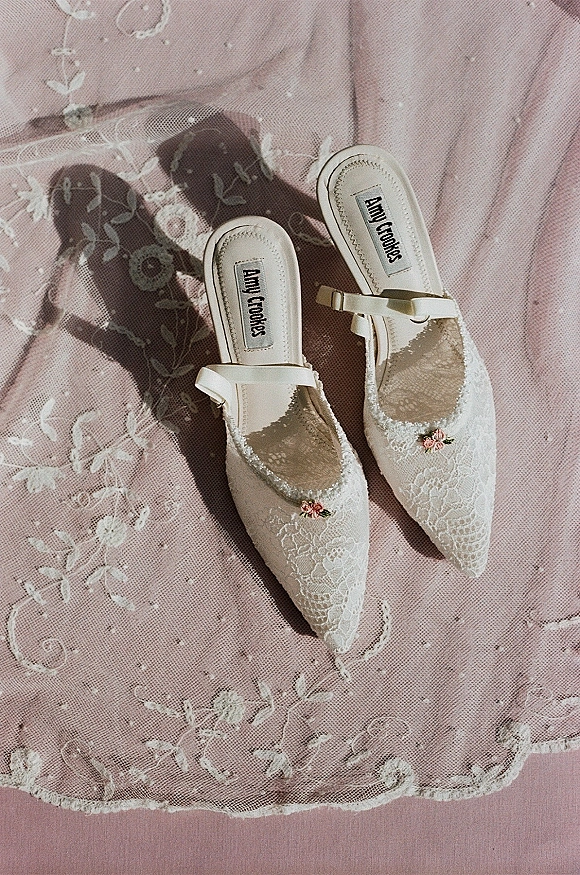 Bridal shoes with lace bridal shoes details, pointed toe mules with ankle straps and floral embroidery, resting on pink fabric backdrop