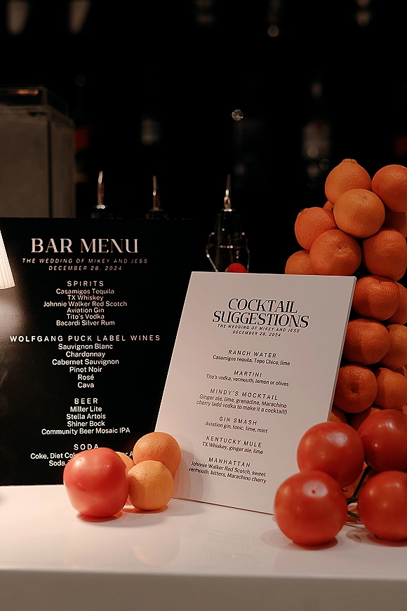 Wedding bar menu and wedding signature cocktails sign displayed in an acrylic holder with oranges and votive candles on a dim bar backdrop