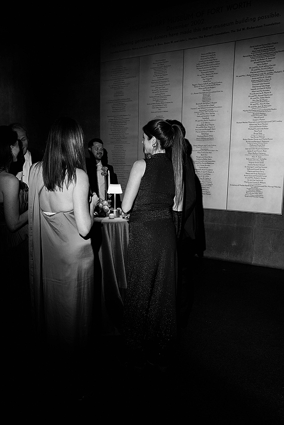 Wedding cocktail hour with wedding guests mingling around a high-top table with a small lamp and candle in a dim indoor venue