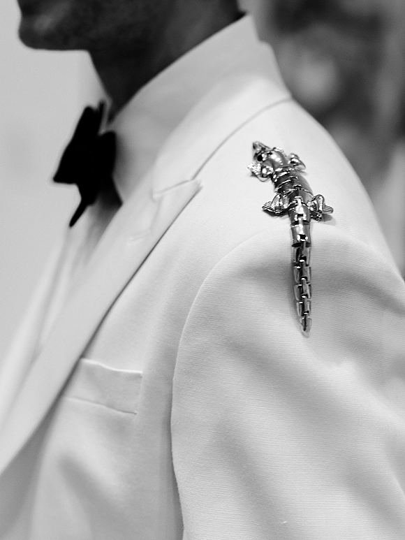 Groom accessory with a lizard boutonniere pin on a white tuxedo jacket, paired with a black bow tie against a softly blurred indoor backdrop
