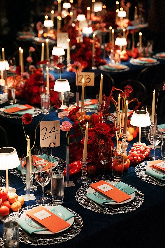 Reception tablescape wedding table setting with red floral and fruit centerpieces, taper candles, and teal napkins on a dark blue cloth in a dim room