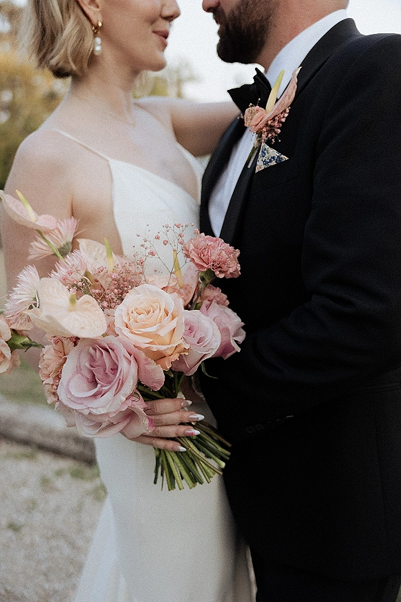 Couple portrait of bride and groom close up holding a blush rose bouquet, groom in black tuxedo and bow tie outdoors by trees