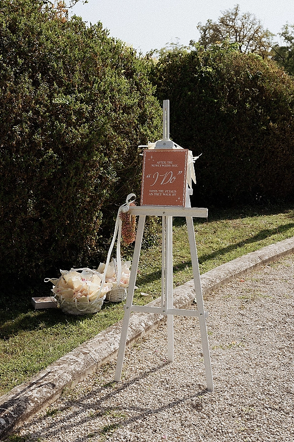 Wedding signage on a white easel with ribbons beside flower petal baskets on a sunny garden path and lawn, ready for guests