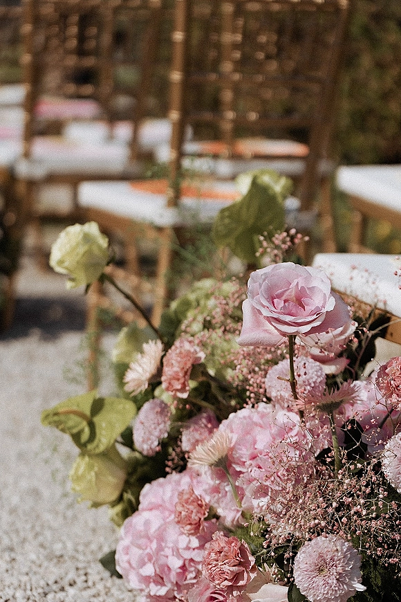 Wedding aisle florals with pink roses and baby's breath on wooden chairs, lining a gravel aisle for an outdoor ceremony setting