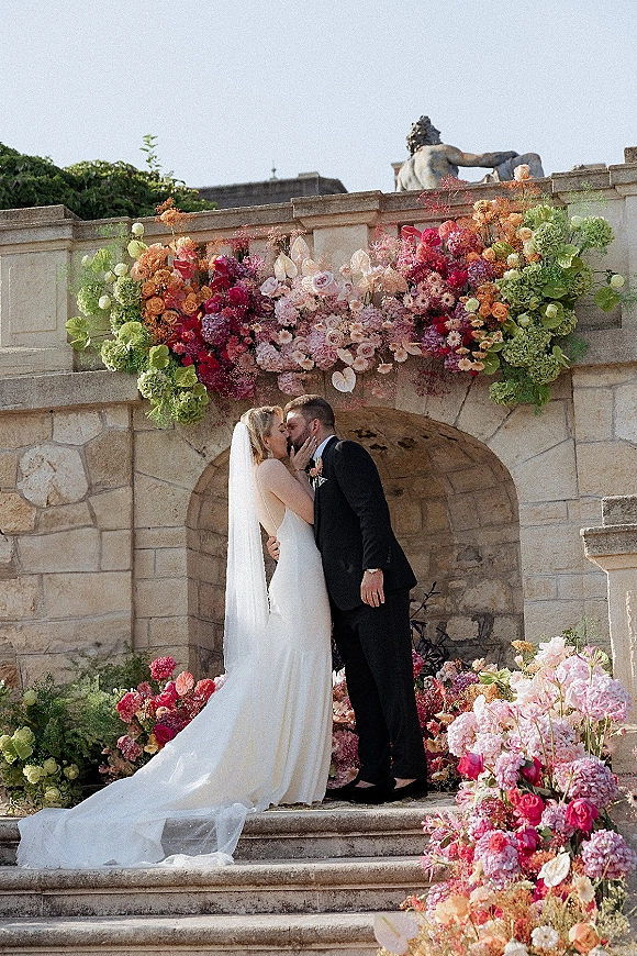 Wedding kiss portrait of bride and groom kissing beneath a floral arch, her veil trailing as they stand on stone steps by a statue
