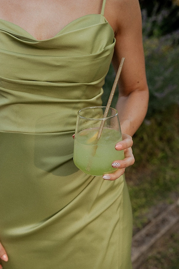 Bridesmaid dress in sage green satin with spaghetti straps and ruched bodice, holding a green cocktail with lemon slice amid garden foliage