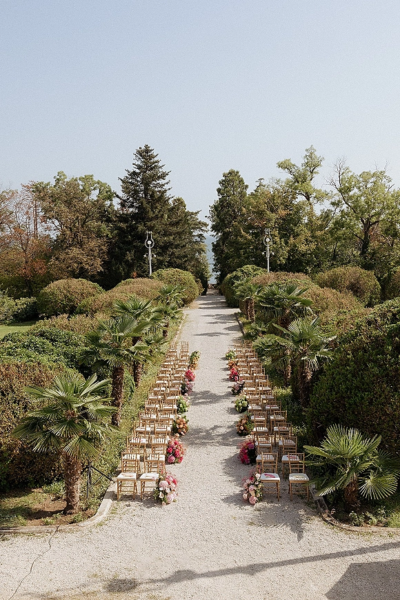 Ceremony aisle design with wood chairs and aisle floral arrangements lining a gravel path in a formal garden with palm trees and hedges