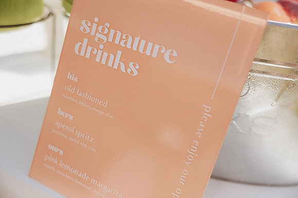 Signature drinks sign with wedding signature drinks menu in white typography on an acrylic sign, displayed on a tabletop beside glassware and dispenser