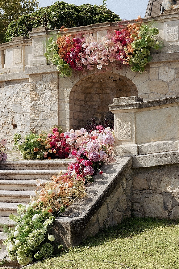 Wedding floral installation with orchids cascading along a stone staircase and archway, blending roses, hydrangea, and greenery outdoors