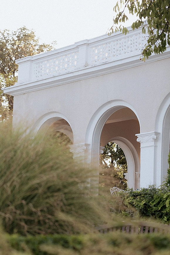 Wedding venue exterior with a white stucco arched colonnade, decorative balustrade, and greenery framing the facade under open sky