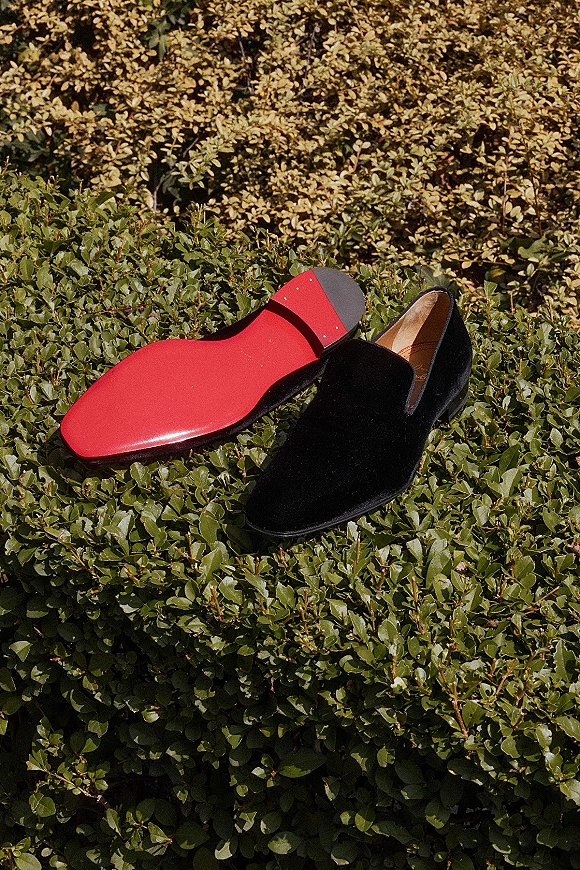 Groom shoes, black velvet loafers with red leather soles, posed on green hedge foliage for an elegant wedding day detail shot