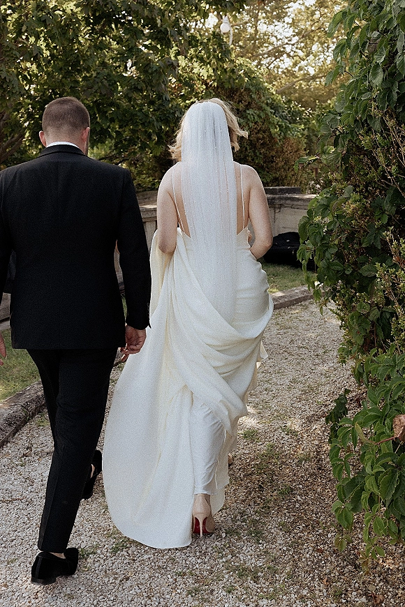 Couple walking away holding hands, bride and groom walking on a gravel garden path, her cathedral veil and dress train flowing behind