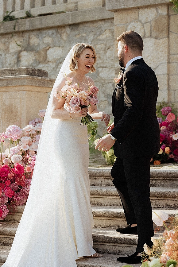 First look moment as bride holding bouquet and groom in tuxedo laugh on stone steps beside a stone wall and garden florals.