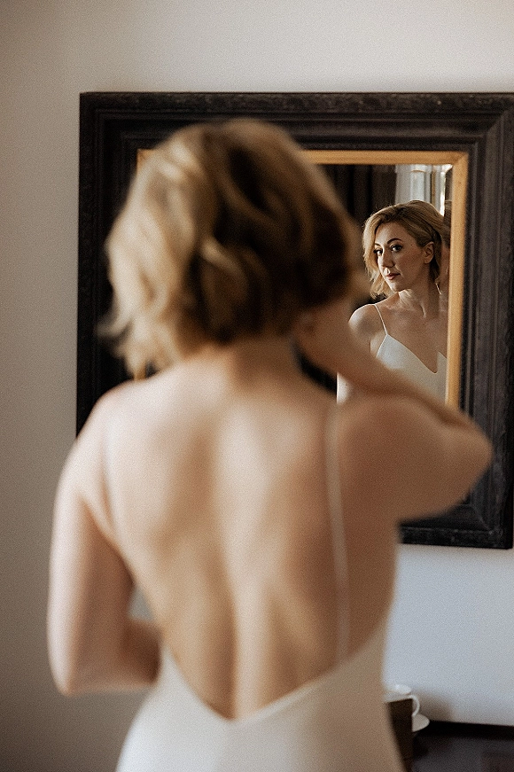 Bridal portrait of a bride in mirror adjusting her hair, wearing a spaghetti-strap low back gown in a minimalist room with dresser and curtain