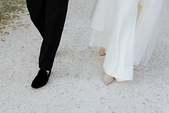 Wedding shoes as bride’s pointed-toe heels and groom black loafers step together on gravel, dress hem and veil brushing near their feet