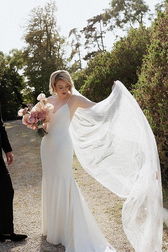 Bridal portrait of a bride holding bouquet, her long veil blowing in the wind along a gravel garden path with hedges and trees