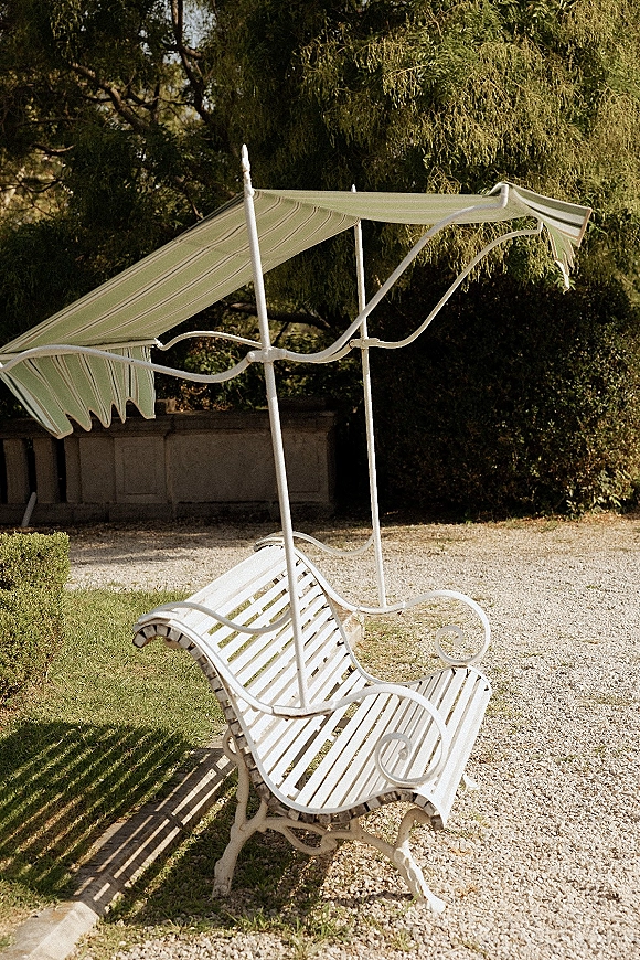 Outdoor lounge seating for wedding cocktail hour with a vintage wrought iron bench under a striped canopy beside hedges and a stone wall