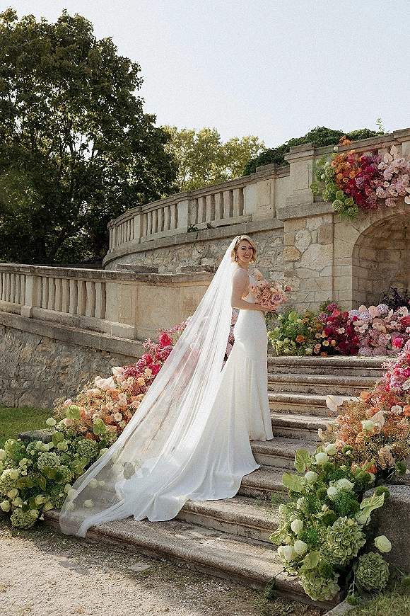 Bridal portrait of a bride on stone stairs in a strapless gown, holding a pastel bouquet with a long cathedral veil by florals